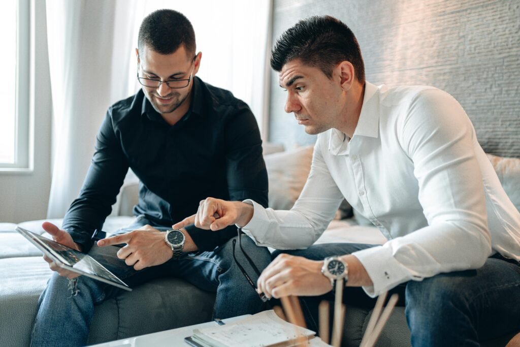 Two businessmen in casual clothing discuss a project on a tablet indoors.